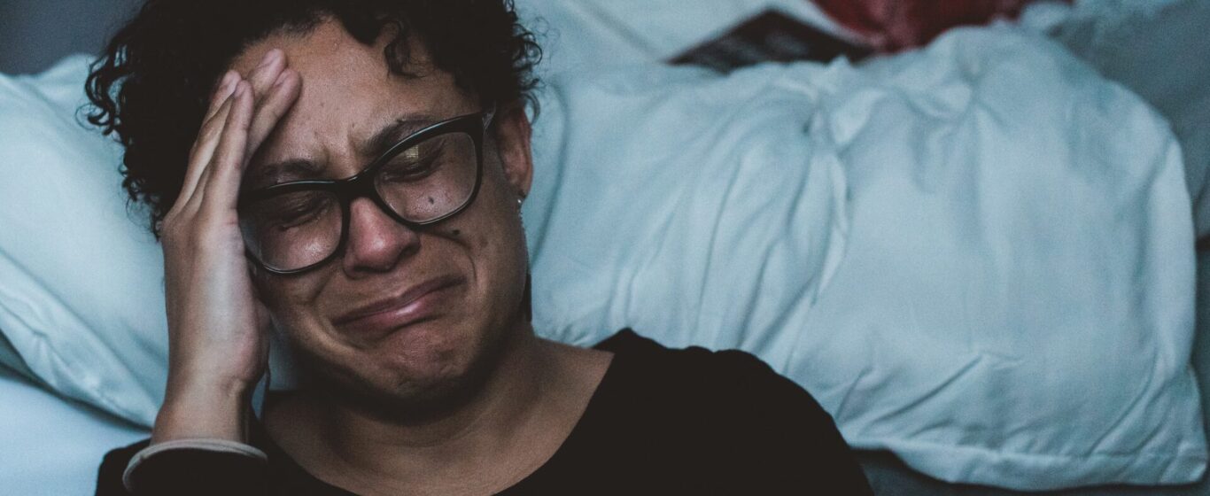 woman wearing glasses crying while sitting on the floor next to her bed